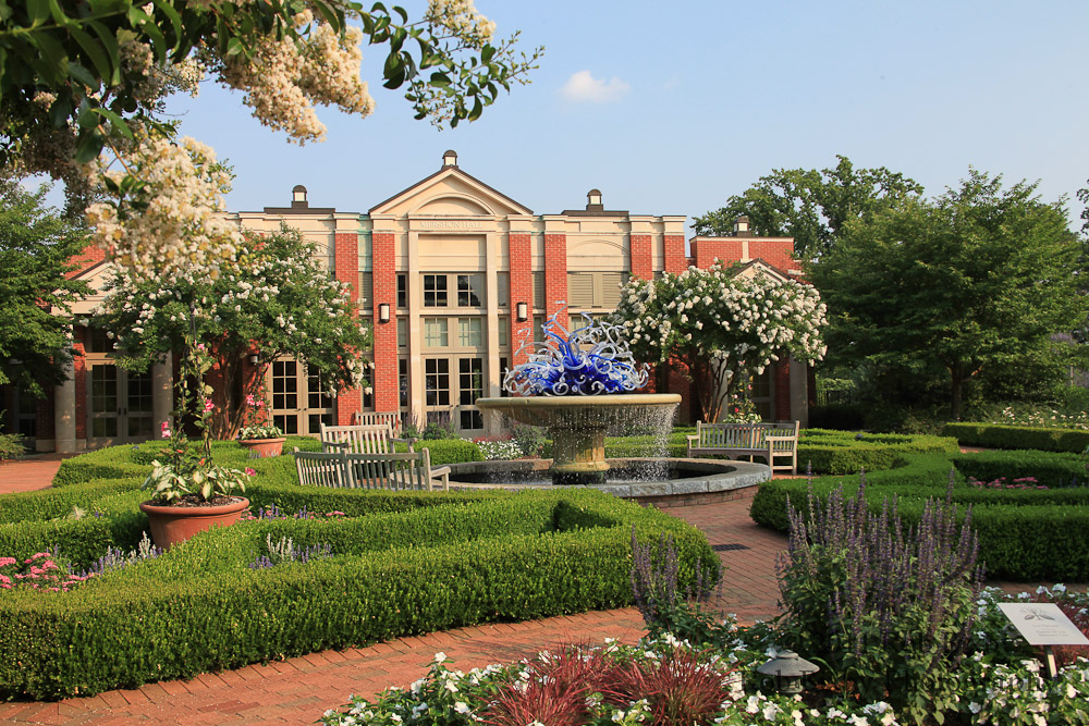 Explore the outdoor garden with a central fountain surrounded by well-maintained hedges and flower beds. A brick building with large windows stands in the background, reminiscent of the Atlanta Botanical Garden experience led by knowledgeable tour guides.