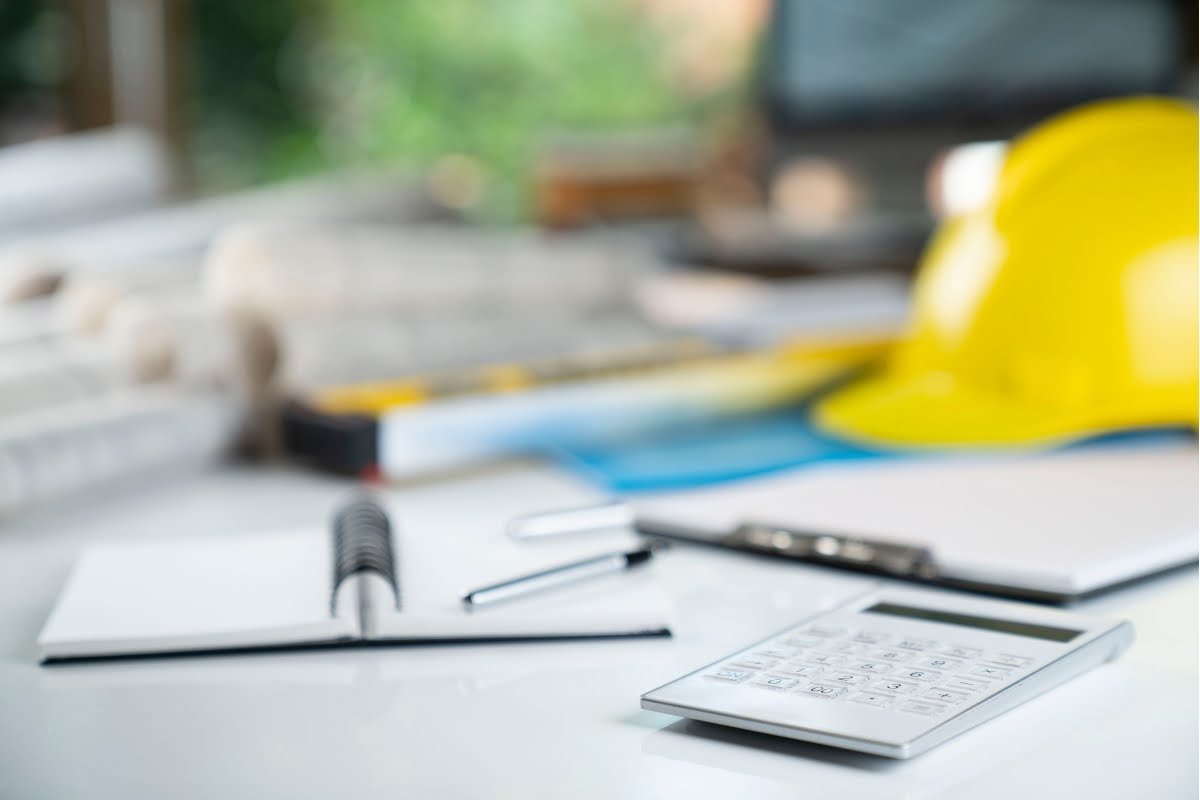 Close-up of a desk with a notepad, pen, calculator, rolled-up blueprints, and a yellow hard hat.