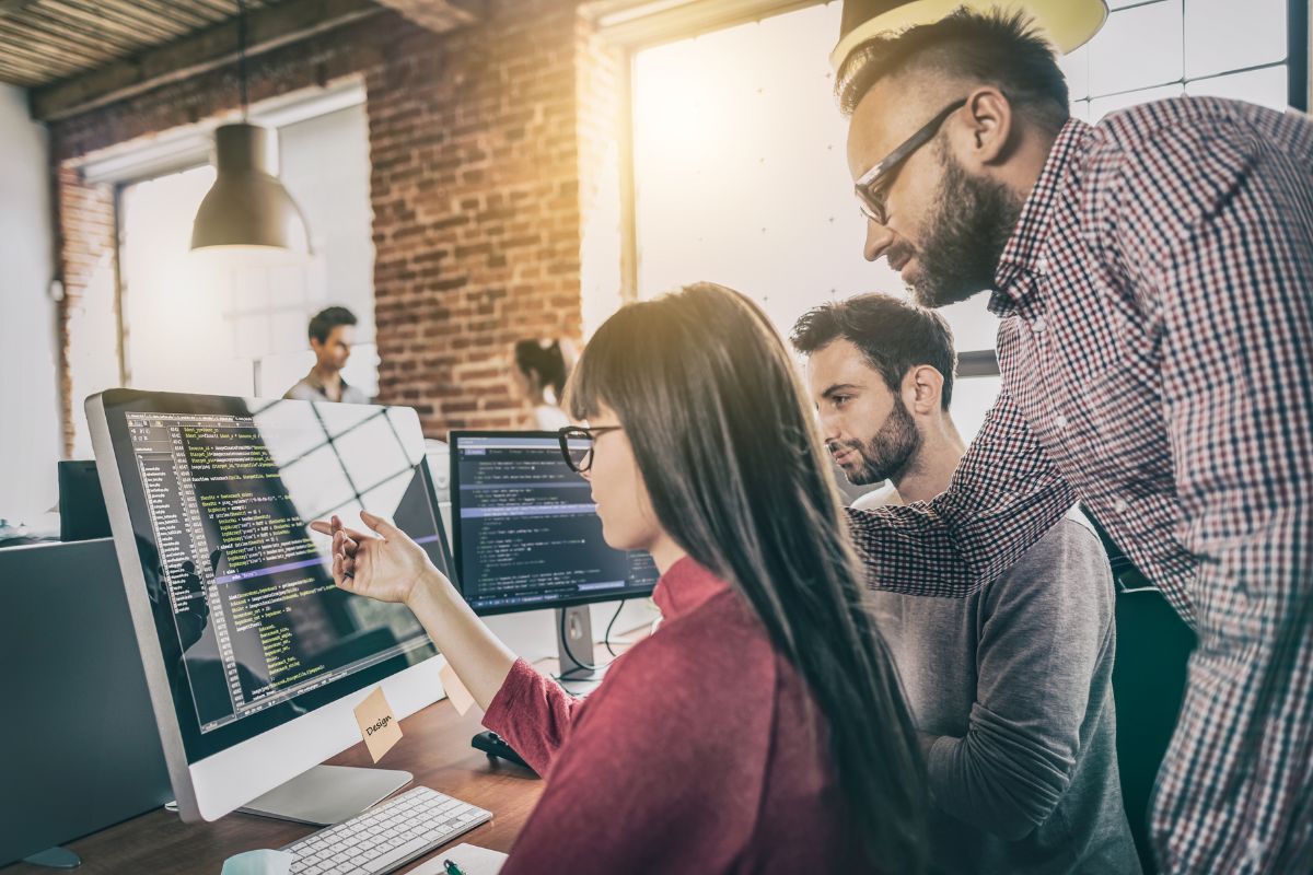 Three people work together at a desk with computers, analyzing code on a monitor in a modern office adorned with exposed brick walls and large windows. They incorporate AI SEO tools to optimize their workflow and enhance their digital strategies.