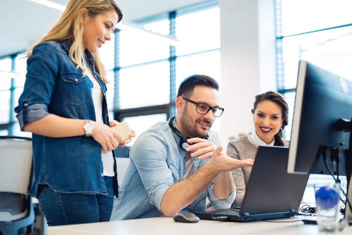 Three people are gathered around a desk with a computer, discussing something on the screen. The man sitting at the desk is gesturing, while the two women stand next to him, engaged in the conversation about AI SEO tools.