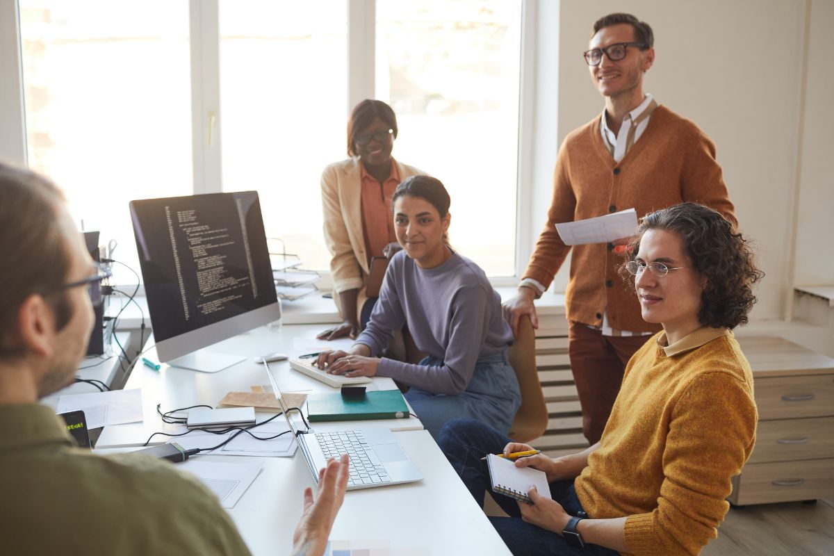 Five people in a well-lit office gather around a desk with a computer, discussing AI SEO tools. The group looks engaged, with one person gesturing while others take notes and listen attentively.