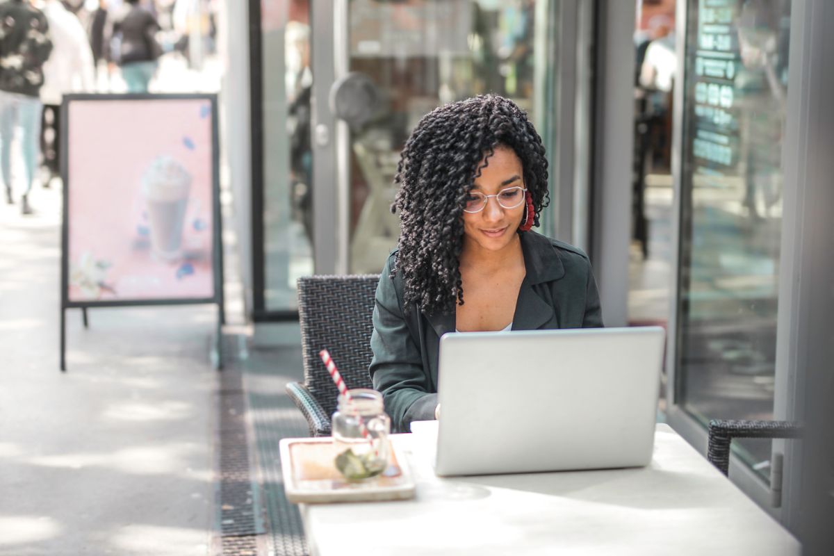 A person with curly hair and glasses is working on a laptop at an outdoor cafe table, possibly using AI SEO tools. There is a beverage with a straw and a small dish on the table. A menu board is visible in the background.