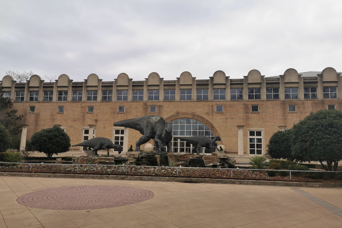 A brick building with arched windows and doors is fronted by life-sized dinosaur statues surrounded by shrubs and a landscaped area, reminiscent of the Atlanta Museum of Natural History.