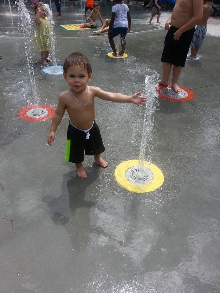 Young children play in an outdoor water fountain area. A shirtless boy in swim shorts stands on a yellow circle, touching one of the water jets, creating unforgettable memories. Other kids are in the background, enjoying their day near the Atlanta Zoo.