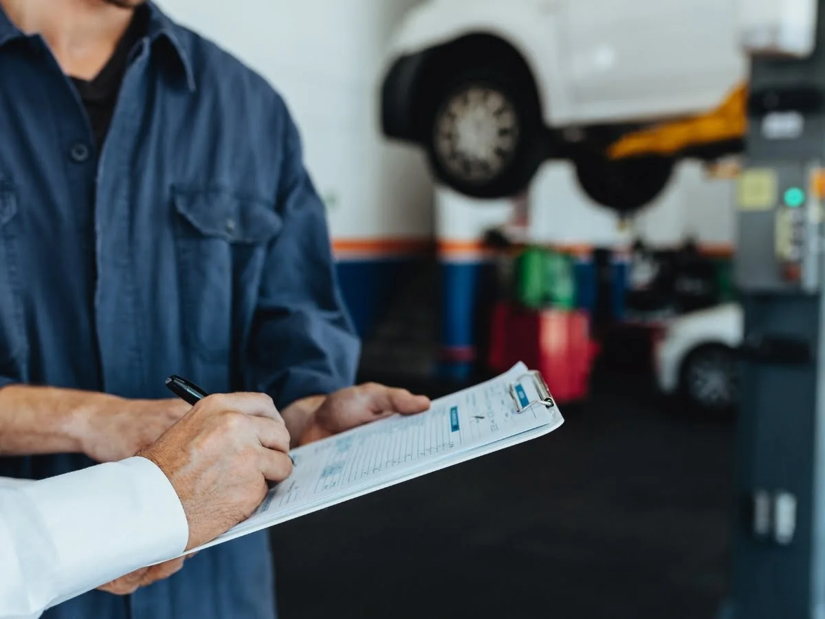 A person in a blue uniform is examining a clipboard while another person writes on it in an auto repair shop with a vehicle elevated on a lift in the background.