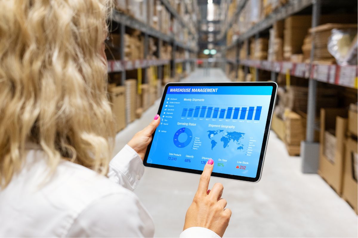 A person in a warehouse holds a tablet displaying automated data collection for warehouse management, including graphs and charts on shipments, operating costs, and shipment geography.