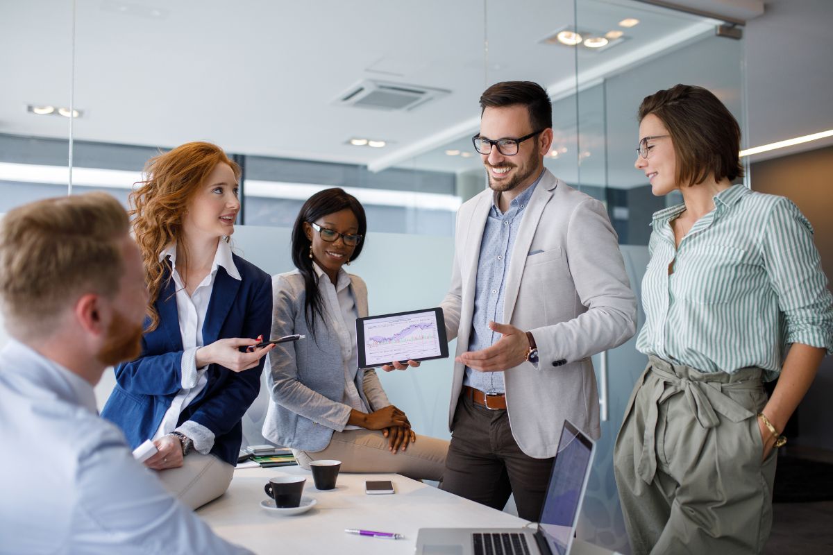 A group of five professionals in a meeting room, with one man standing and holding a tablet displaying a chart on CRM integration, while the others listen and engage in discussion.