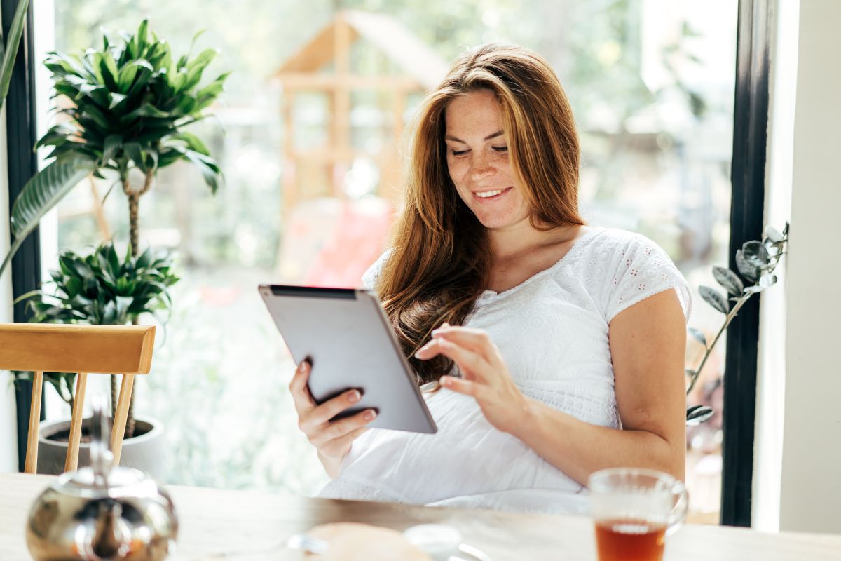 A woman sits at a table by a window, using a tablet. There is a glass of tea on the table and some plants in the background. She reviews SEO strategies, focusing on optimizing robots.txt files.