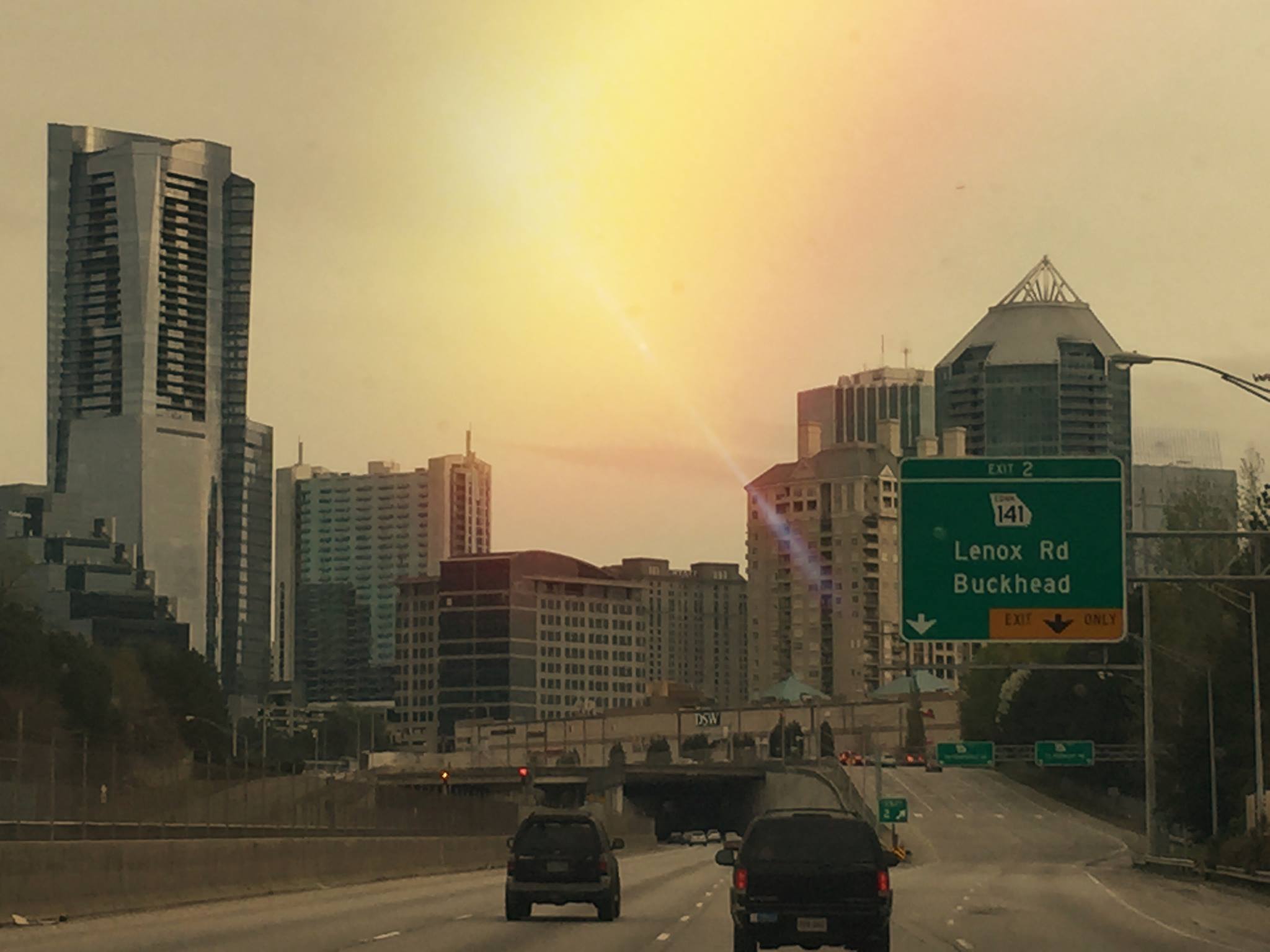 View from a car driving on an urban highway, passing tall buildings in a city with a green road sign indicating the exits for Lenox Rd and Buckhead. The sun is shining brightly in the sky as you approach the vibrant Buckhead Atlanta shopping district.