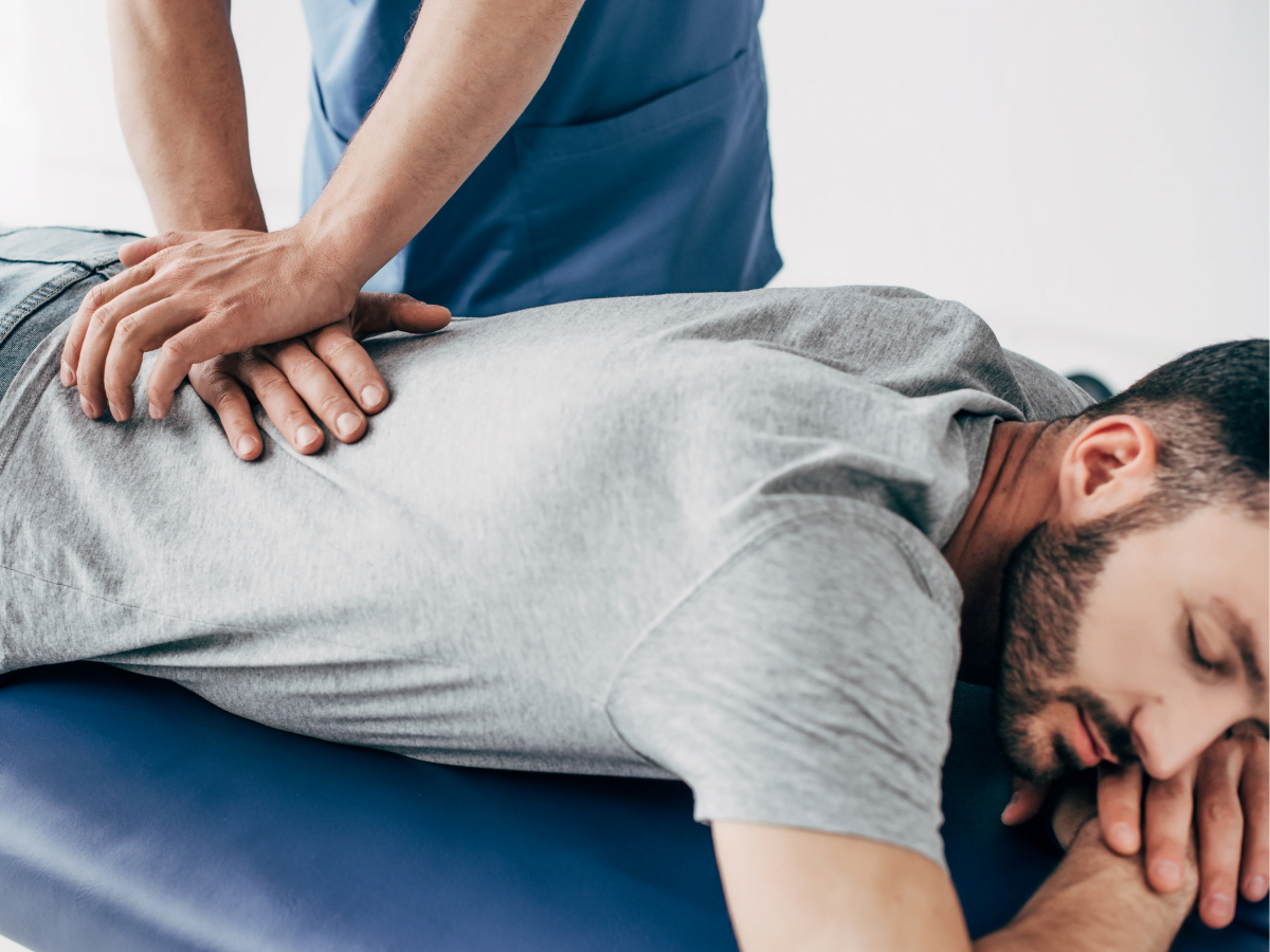 A person in a gray shirt lies face down on a blue table while another person performs a back massage.