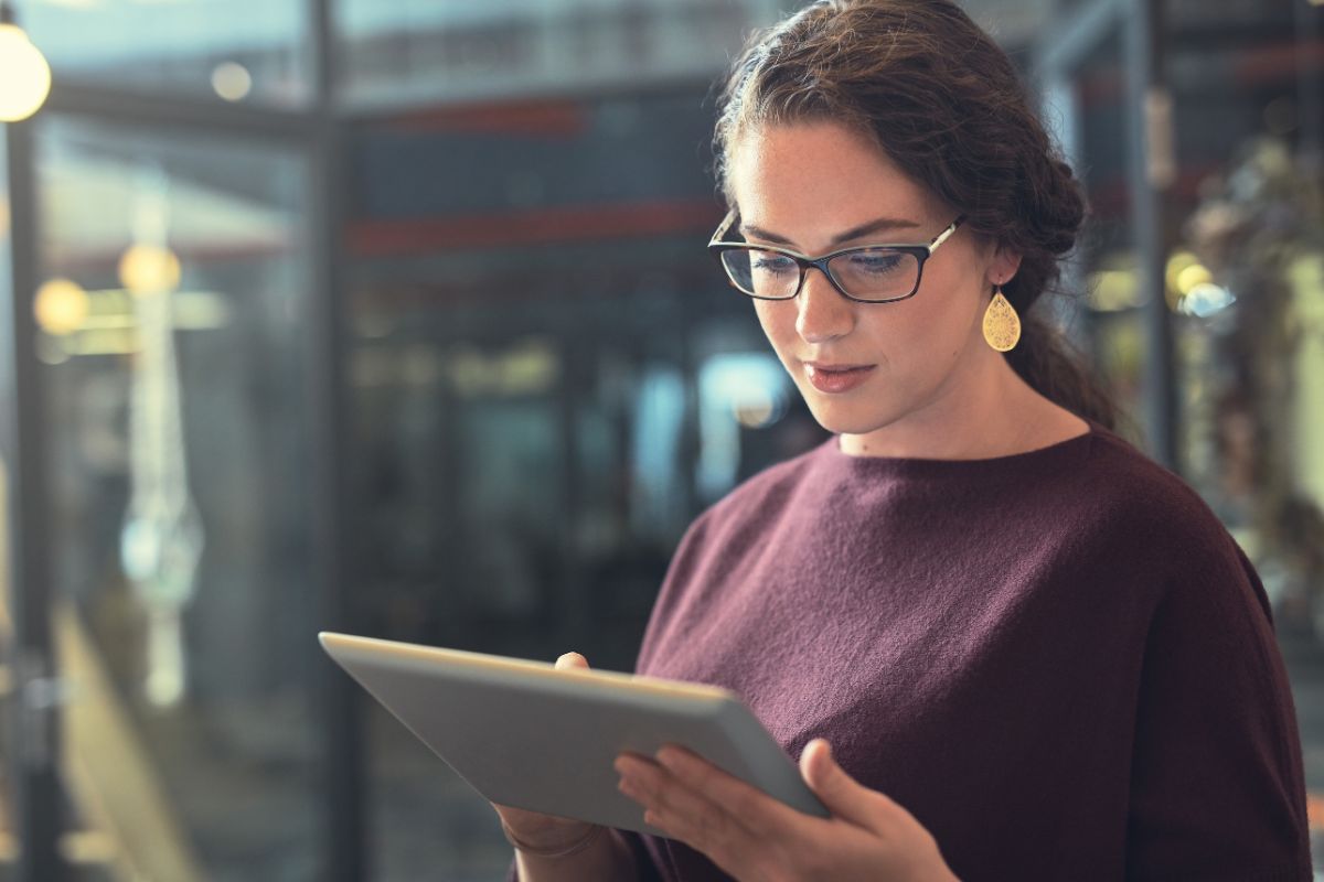 A woman wearing glasses and earrings is working on a tablet in an indoor setting with glass walls in the background, seamlessly integrating email marketing automation tools into her workflow.