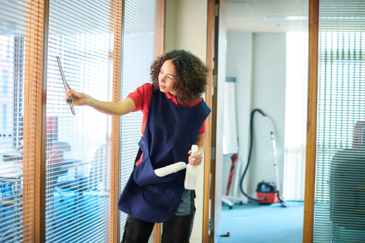 Person with curly hair cleaning office windows with a squeegee and spray bottle, wearing a blue apron over a red shirt. Vacuum cleaner and cleaning supplies are visible in the background.