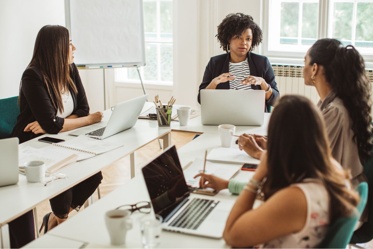 Four women are seated at a conference table with laptops, notebooks, and coffee cups, engaged in a meeting about optimizing their website. One woman is speaking about the importance of robots.txt for SEO while others listen attentively.