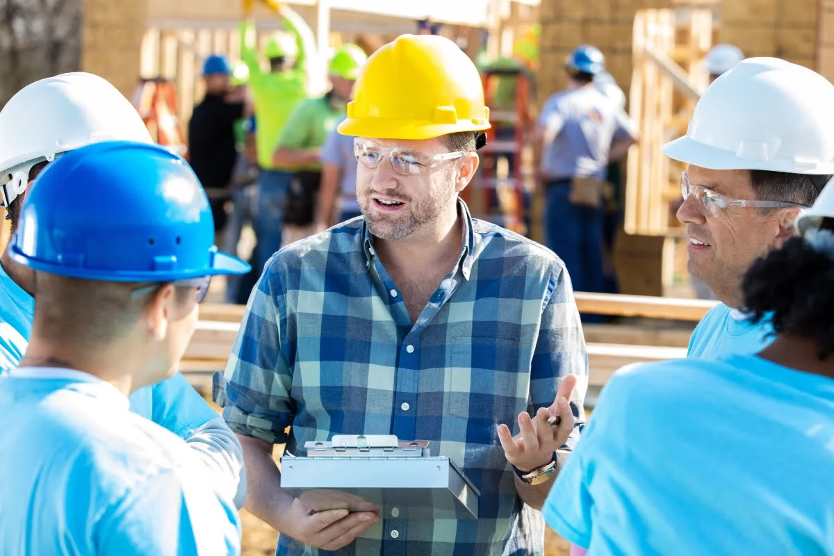 A construction manager in a yellow hard hat holds a clipboard and speaks to a group of workers wearing blue hard hats at a building site. Other workers and construction activities are visible in the background.