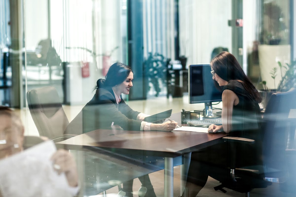 Two women are sitting at a desk in a modern office, one of them is showing a document to the other. They appear to be engaged in a discussion.