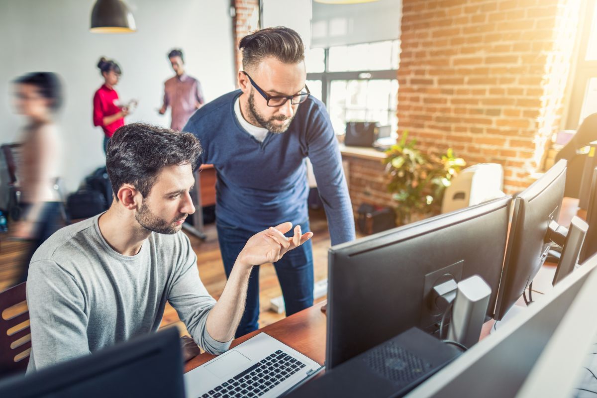 Two men are working on a computer in an office with colleagues in the background. The seated man is gesturing towards the screen, likely discussing CRM for business automation, while the standing man attentively looks at it.