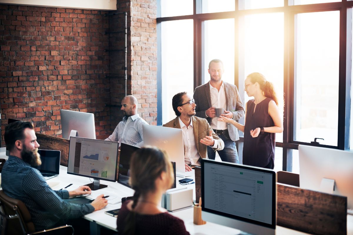 A group of people are working in a modern office with large windows and exposed brick walls. Some are seated at desks with computers, focusing on CRM integration, while others are standing and conversing about their projects.