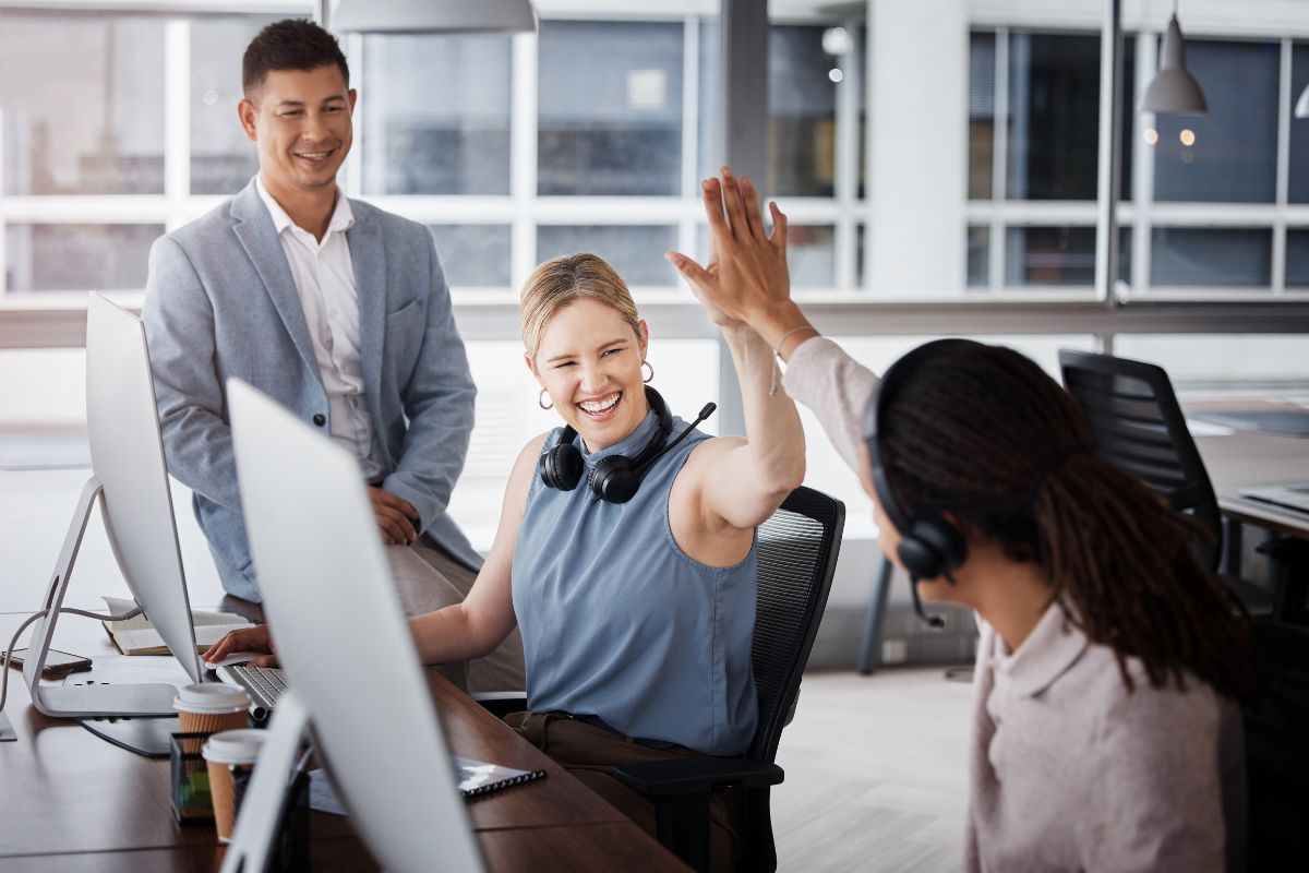 Three colleagues in an office setting, two seated and one standing. The seated woman and man are giving a high-five, smiling as they celebrate a successful CRM marketing automation implementation. Computers and office supplies are visible on the desks.