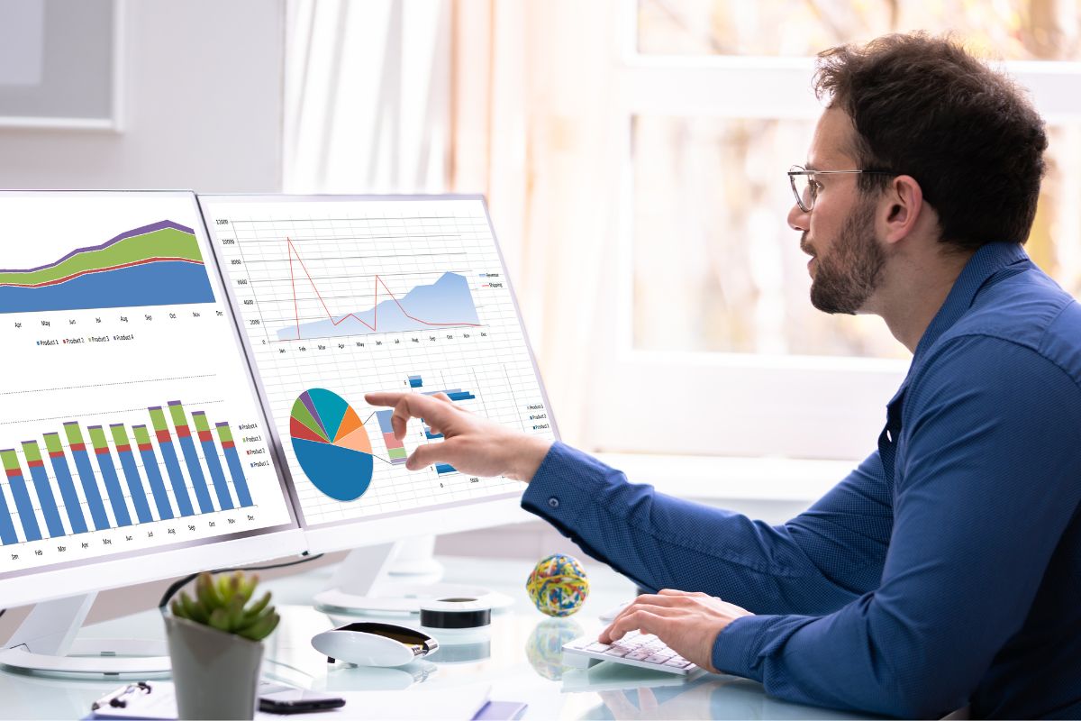 A man in a blue shirt analyzes data on two computer monitors displaying various charts and graphs in an office setting, exploring ways to automate data collection. A small plant and a decorative ball are on the desk.