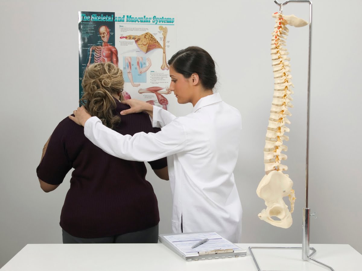 A healthcare professional in a white coat examines a patient's back in a medical office with an anatomical spine model and a skeletal system poster in the background.