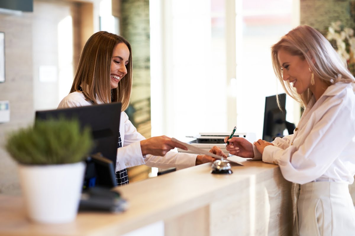 A receptionist hands a document to a woman who is signing it at a reception desk using spa management software.