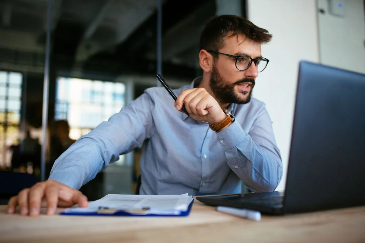 A man wearing glasses sits at a desk, holding a pen in one hand and looking at a laptop screen, possibly researching duplicate content in SEO. A clipboard with papers is on the desk.