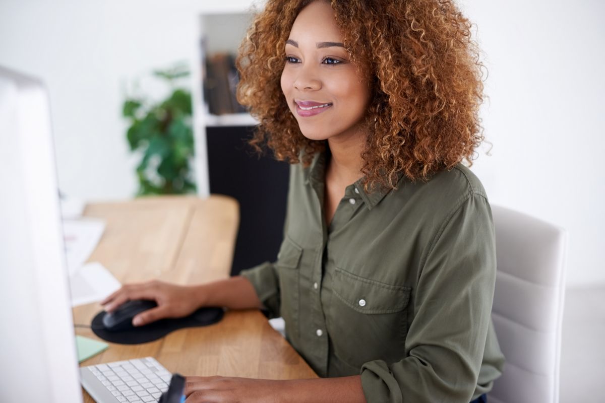 A woman with curly hair, wearing a green shirt, uses a computer mouse and keyboard while smiling at her monitor, as she navigates through email marketing automation tools.