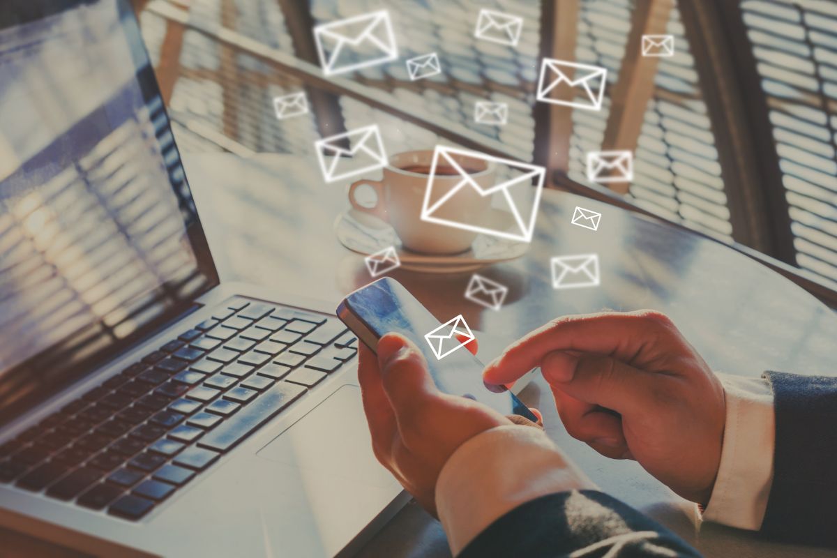 Person using a smartphone next to a laptop on a table, with email marketing automation tools icons floating above the phone.