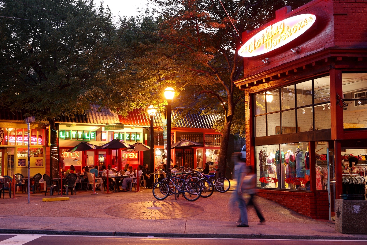 Street scene at dusk with illuminated storefronts, including a pizzeria and a clothing store in Bohemian Atlanta's Little Five Points Shopping district. Bicycles are parked near outdoor seating while pedestrians walk past. Trees and lampposts line the sidewalk.