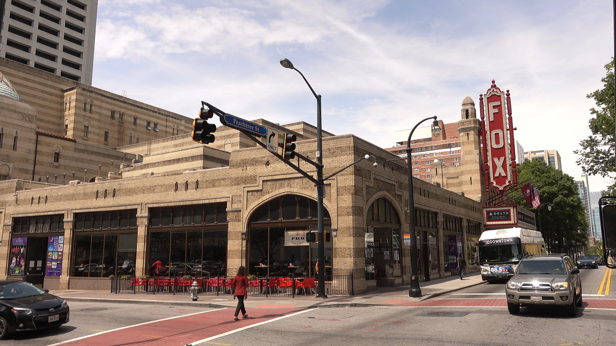 Street view of the Fox Theatre in Atlanta, prominently featuring its marquee. Nearby pedestrians walk casually, and cars remain stopped at a traffic light during the day.