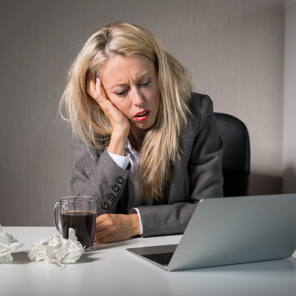 A tired person with long blonde hair looks at a laptop, head resting on one hand. A mug and crumpled tissues are on the desk, likely evidence of late nights spent implementing business automation or managing a small business CRM.