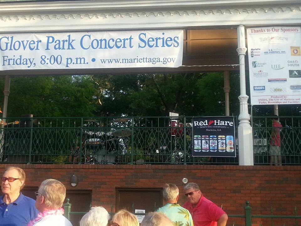 People gather in front of a stage at the Glover Park Concert Series event in Marietta, with banners displaying sponsor logos and event information. This musical gem brings the community together for an unforgettable evening.