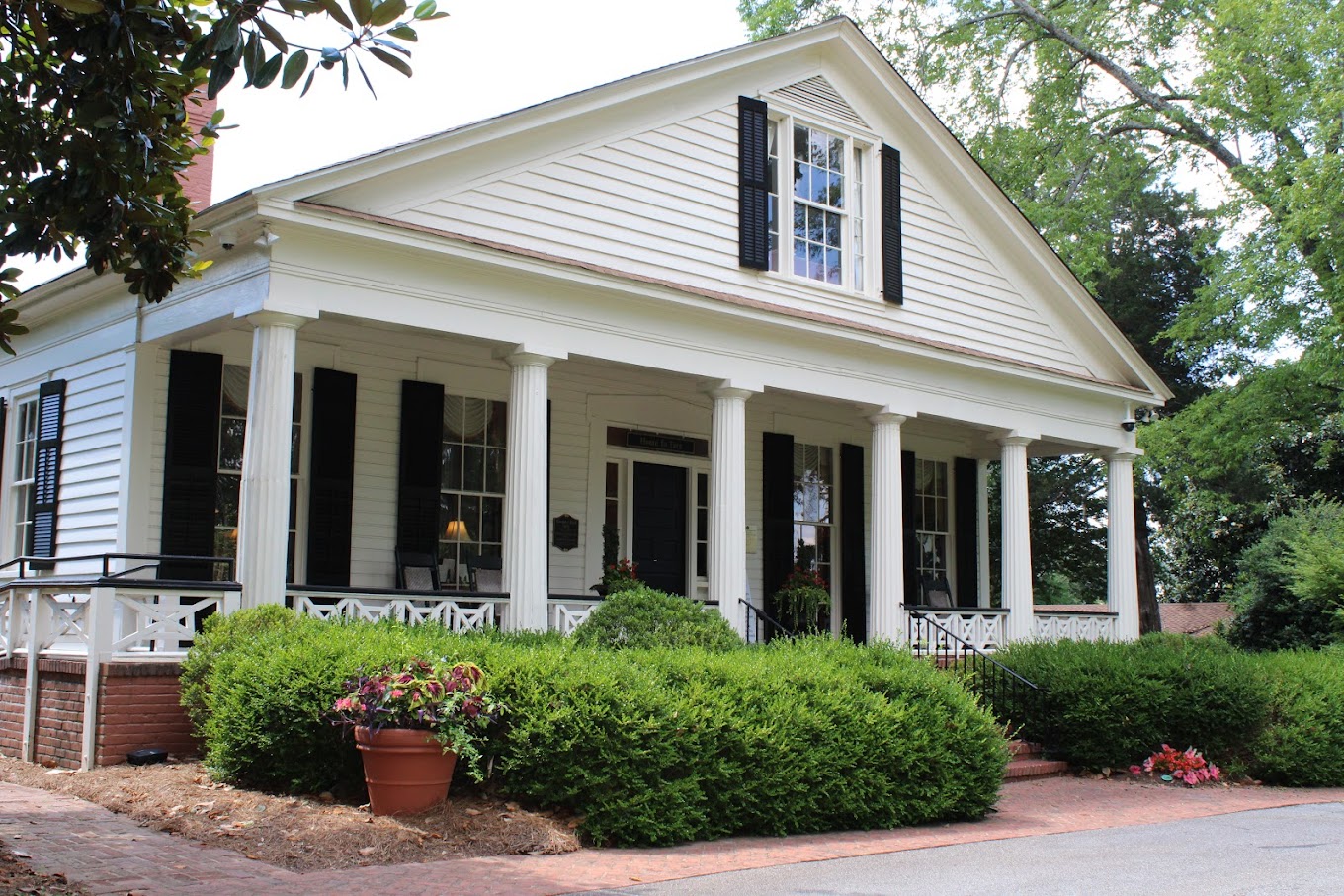 A white, single-story house with black shutters and a brick foundation is surrounded by bushes and trees. Reminiscent of scenes from the Gone With the Wind Museum in Marietta, it features a wide front porch supported by columns and a potted plant by the entrance.