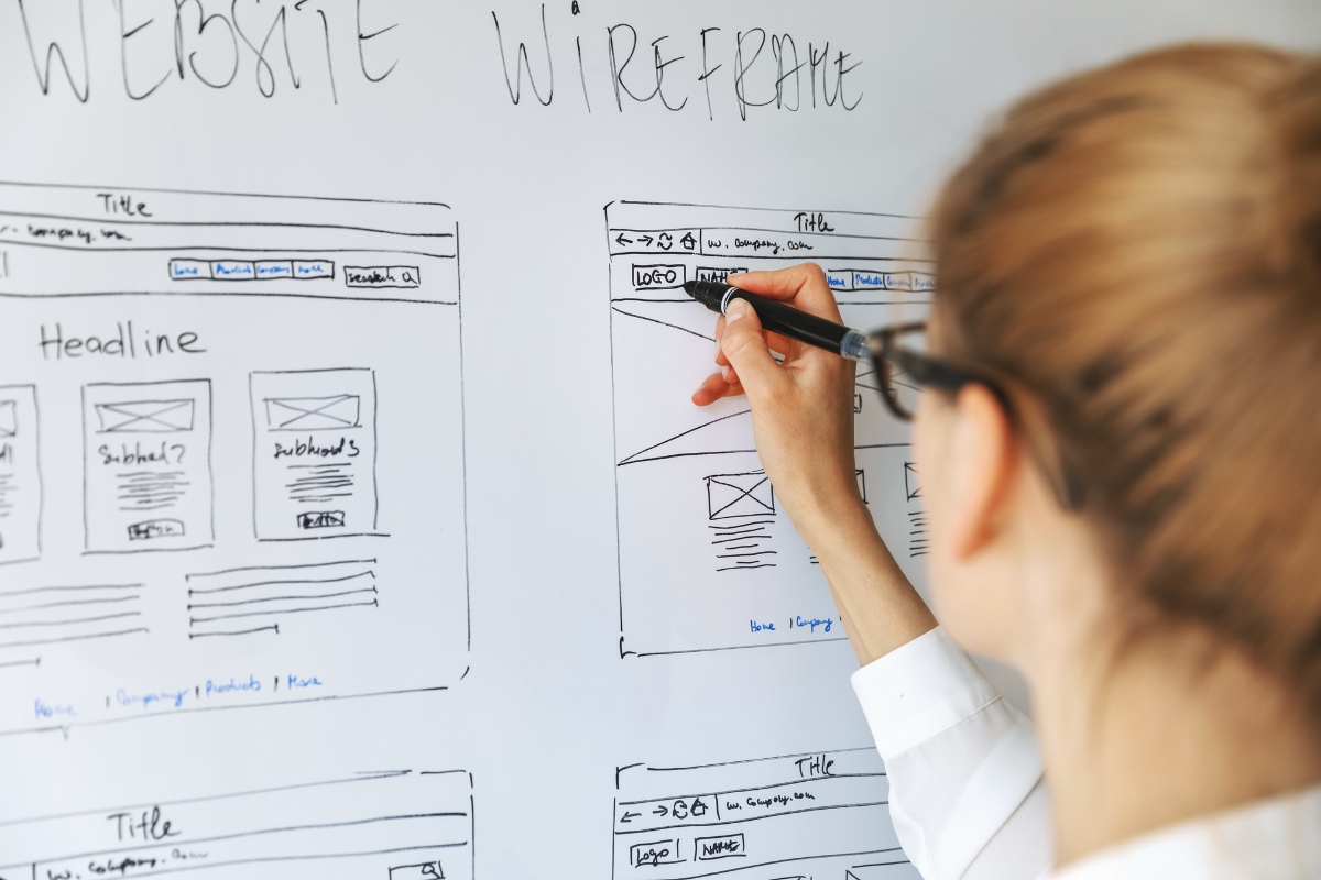 A person with glasses is drawing website wireframes on a whiteboard, using a black marker to create outlines for different web page sections such as titles and headlines, while considering how website design affects sales.