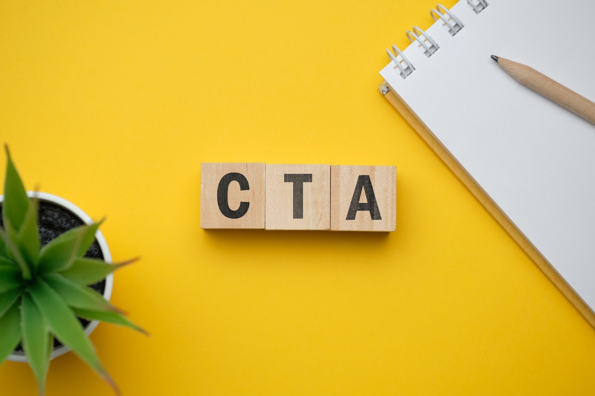 Wooden blocks with "CTA" letters are placed on a yellow background next to a small plant, a notebook, and a pencil, subtly hinting at how website design affects sales through effective call-to-action elements.