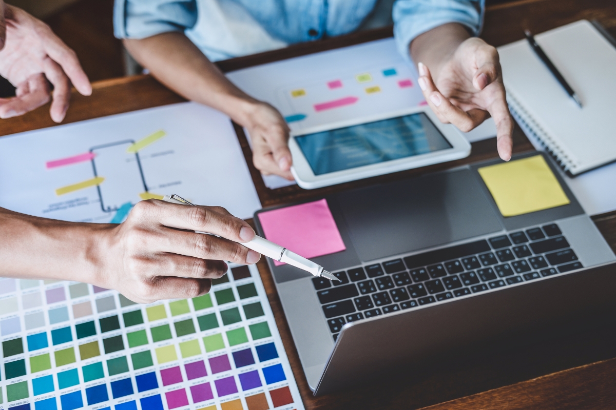 Three people collaborate at a desk with a laptop, a tablet, color charts, sticky notes, and printed diagrams to explore how website design affects sales effectiveness.