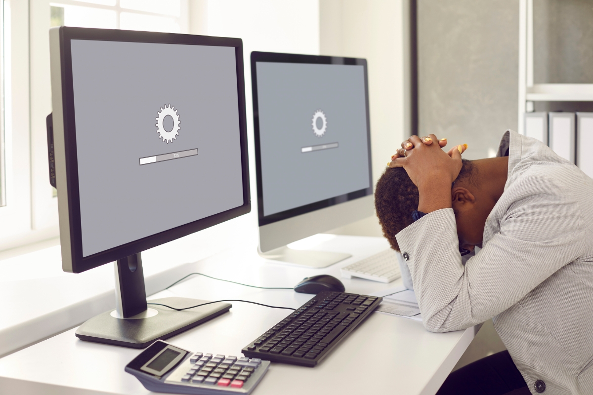 Person sitting at a desk with their head in their hands in frustration, pondering how does website design affect sales. Two computer monitors show a loading screen. A keyboard, mouse, and calculator clutter the desk.