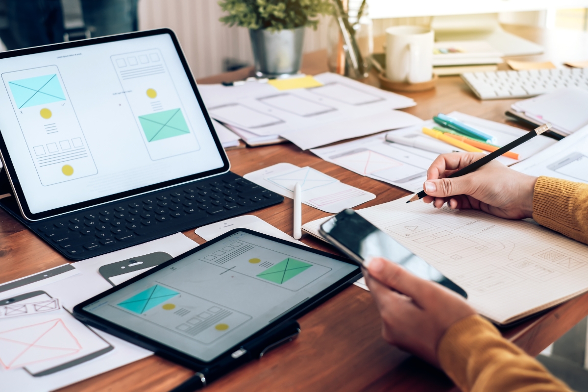 A person working on website design sketches on a paper notebook, pondering how website design affects sales, while referencing an iPad and a tablet displaying wireframes. The workspace is cluttered with various papers and design materials.