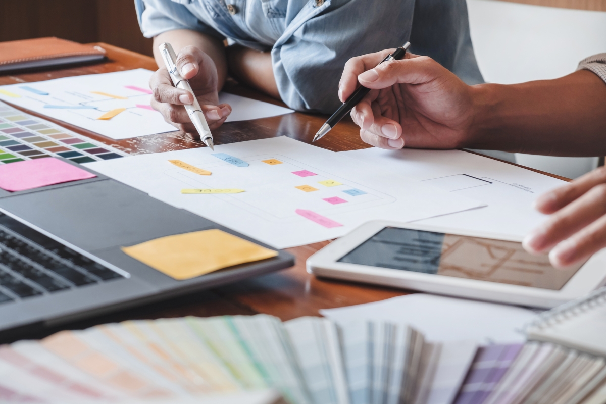 Two people collaboratively working on a project at a desk, using a laptop, tablet, color swatches, and various papers with design sketches and sticky notes, discussing how website design affects sales.