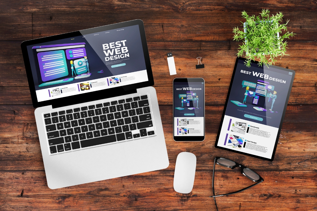 Laptop, smartphone, and tablet displaying a web design website on a wooden desk with a mouse, reading glasses, USB drive, and a small plant—every element meticulously placed to answer the question: how does website design affect sales?