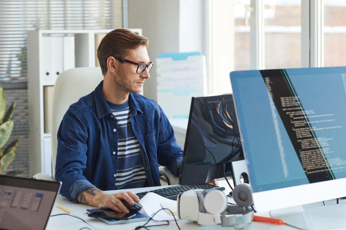 A person with glasses sits at a desk working on two computer monitors displaying code related to CRM integration in a bright office.