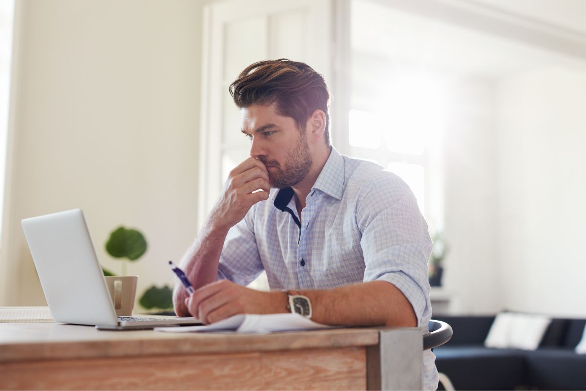 A man in a white shirt sits at a desk, concentrating on a laptop while holding a pen. Papers on duplicate content in SEO are spread out on the desk in front of him.