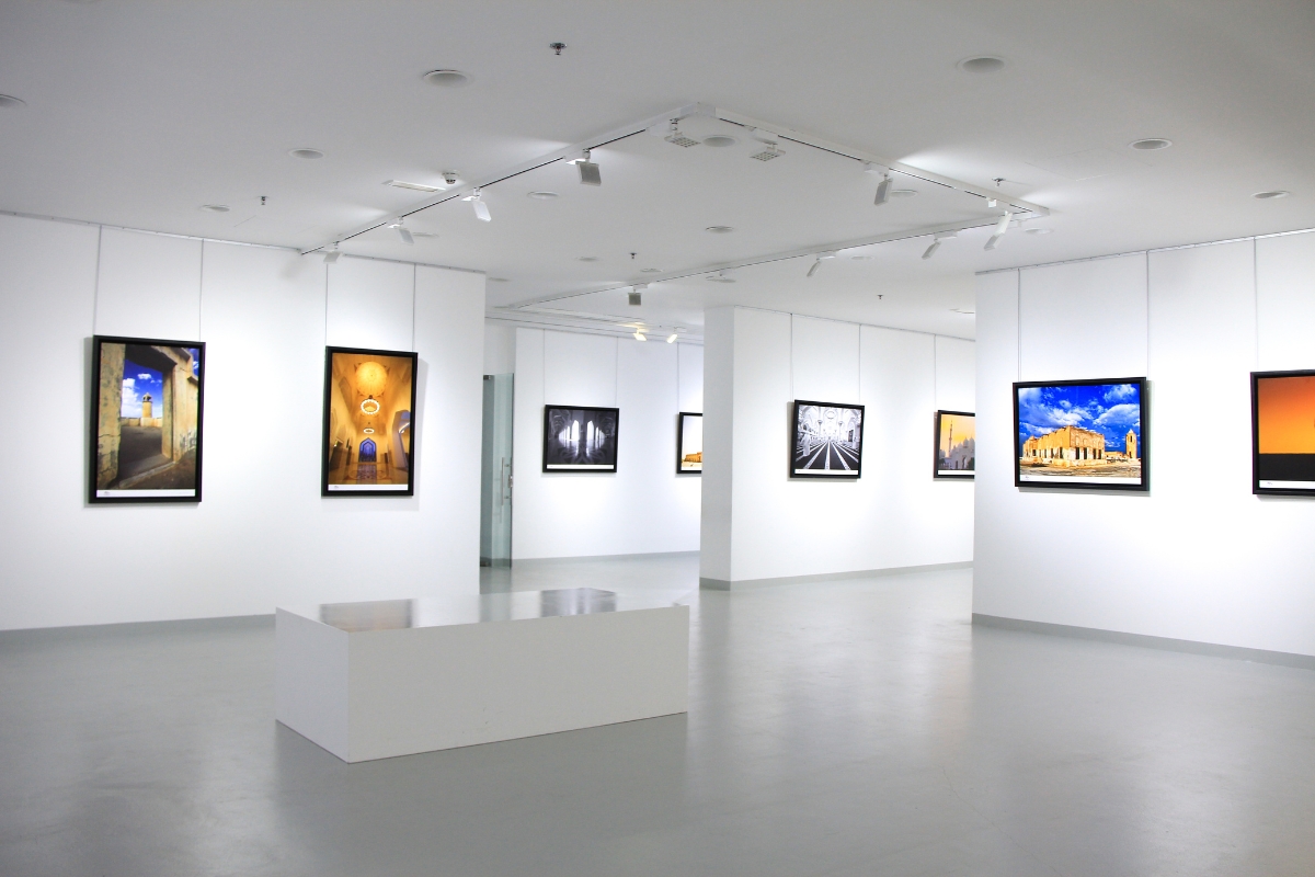 A brightly lit art gallery with pictures hung on white walls, featuring various photographs including landscapes and architectural scenes. A rectangular white bench is placed in the center of the room, creating a serene environment perfect for contemplating both art and how to market your small business.