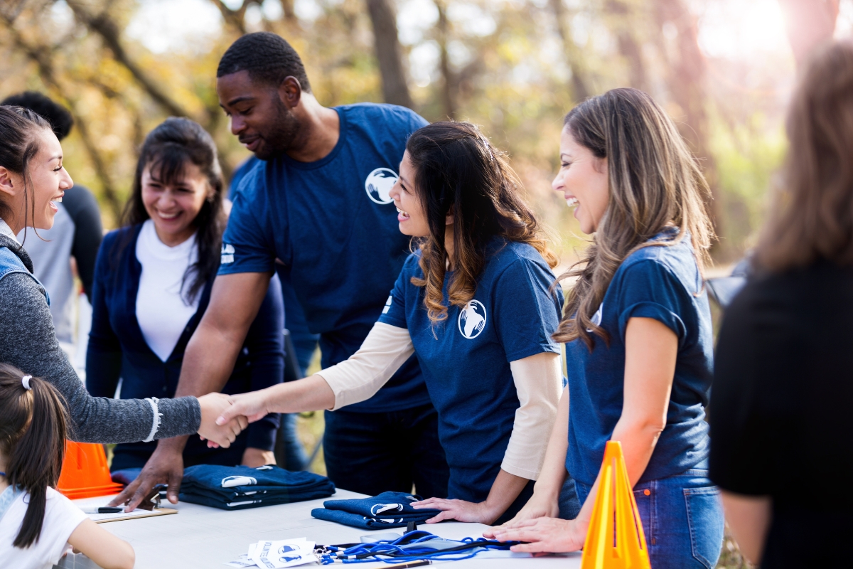 People wearing blue shirts stand behind a table, engaging with attendees at an outdoor event, shaking hands and smiling. The table has various items displayed on it, showcasing how to market your small business effectively.