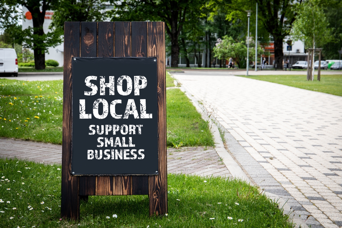 A wooden sign on a grassy area reads "Shop Local, Support Small Business," exemplifying how to market your small business effectively. A sidewalk and trees are visible in the background.