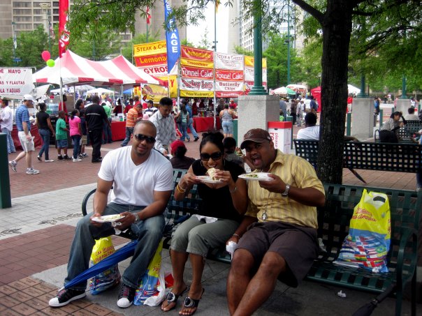 Three people sitting on a bench eating food at Festival Peachtree Latino, surrounded by vibrant activities, food stalls, and other attendees enjoying the attractions in the background.