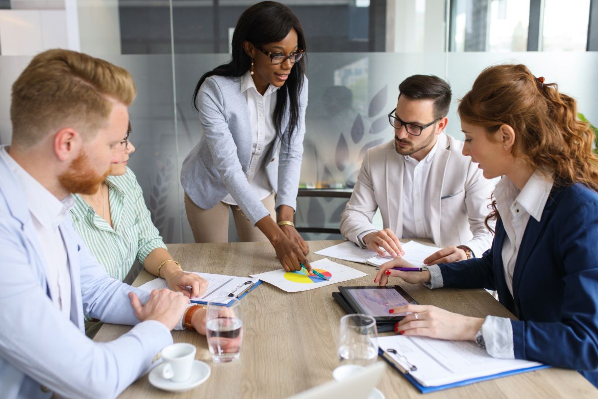 Five business professionals in a meeting room, one standing and pointing at a chart on the table. Others are seated, paying attention and taking notes, discussing lead generation marketing strategies.