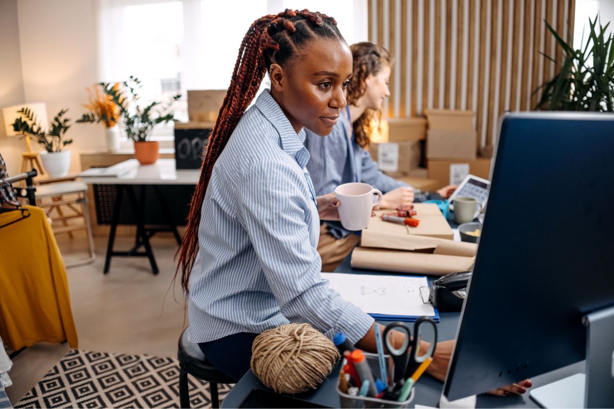 A woman with braided hair holds a mug and works at a computer in an office setting, likely focusing on SEO for multiple locations, with another person working in the background.