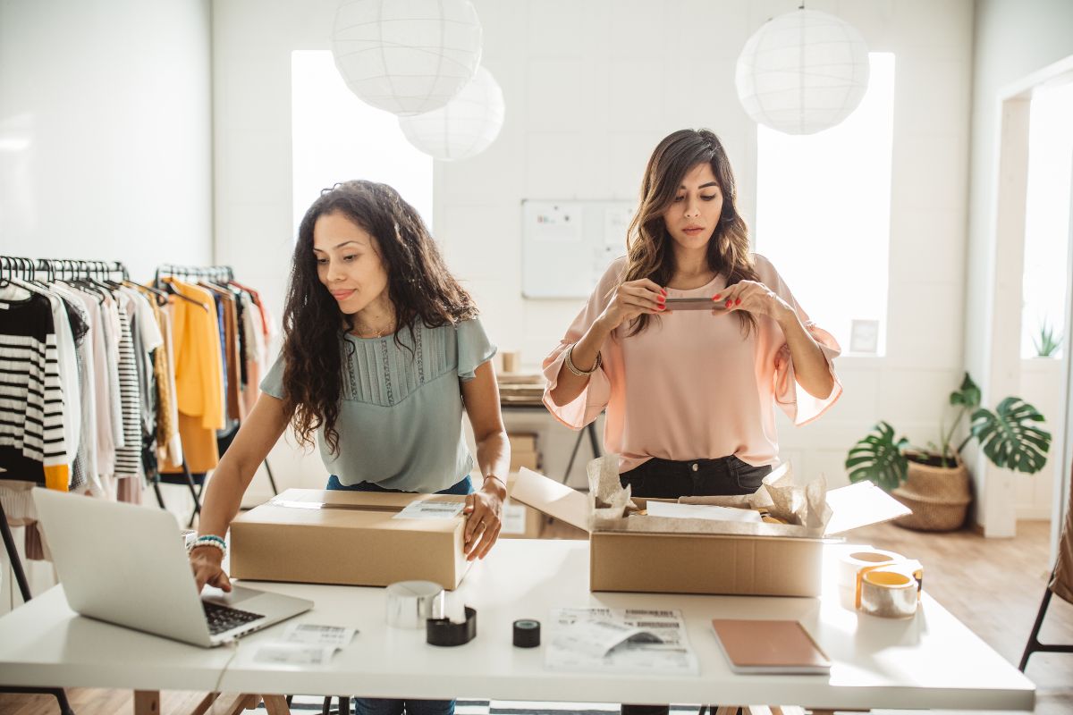 Two women are preparing packages at a table with a laptop, shipping supplies, and a cup of coffee in a well-lit room with clothes hanging in the background, likely discussing their strategy for implementing SEO for multiple locations.