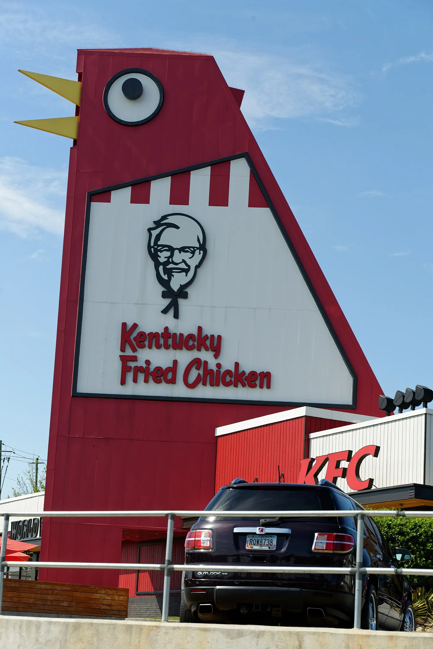 A red building shaped like a chicken with the iconic Kentucky Fried Chicken (KFC) logo stands as a landmark in Marietta, known locally as the Big Chicken, with a car parked in front of it.